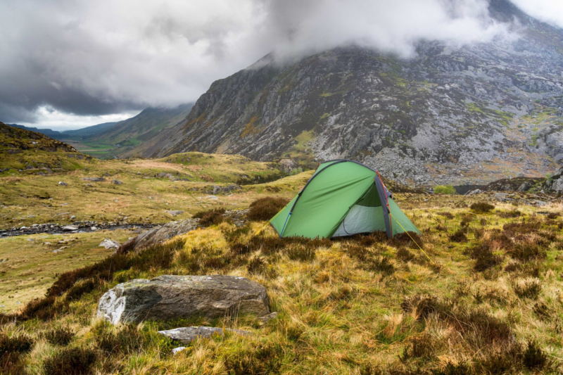 A lone green tent pitched on grassy, heather-studded moorland beside scattered boulders in a steep, rocky mountain valley partly shrouded in low clouds.