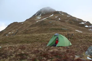 Person in a red jacket and beanie sits at the doorway of a green tent pitched on windswept brown grass with trekking poles beside it, set against a steep mist-capped mountain slope with patches of snow.