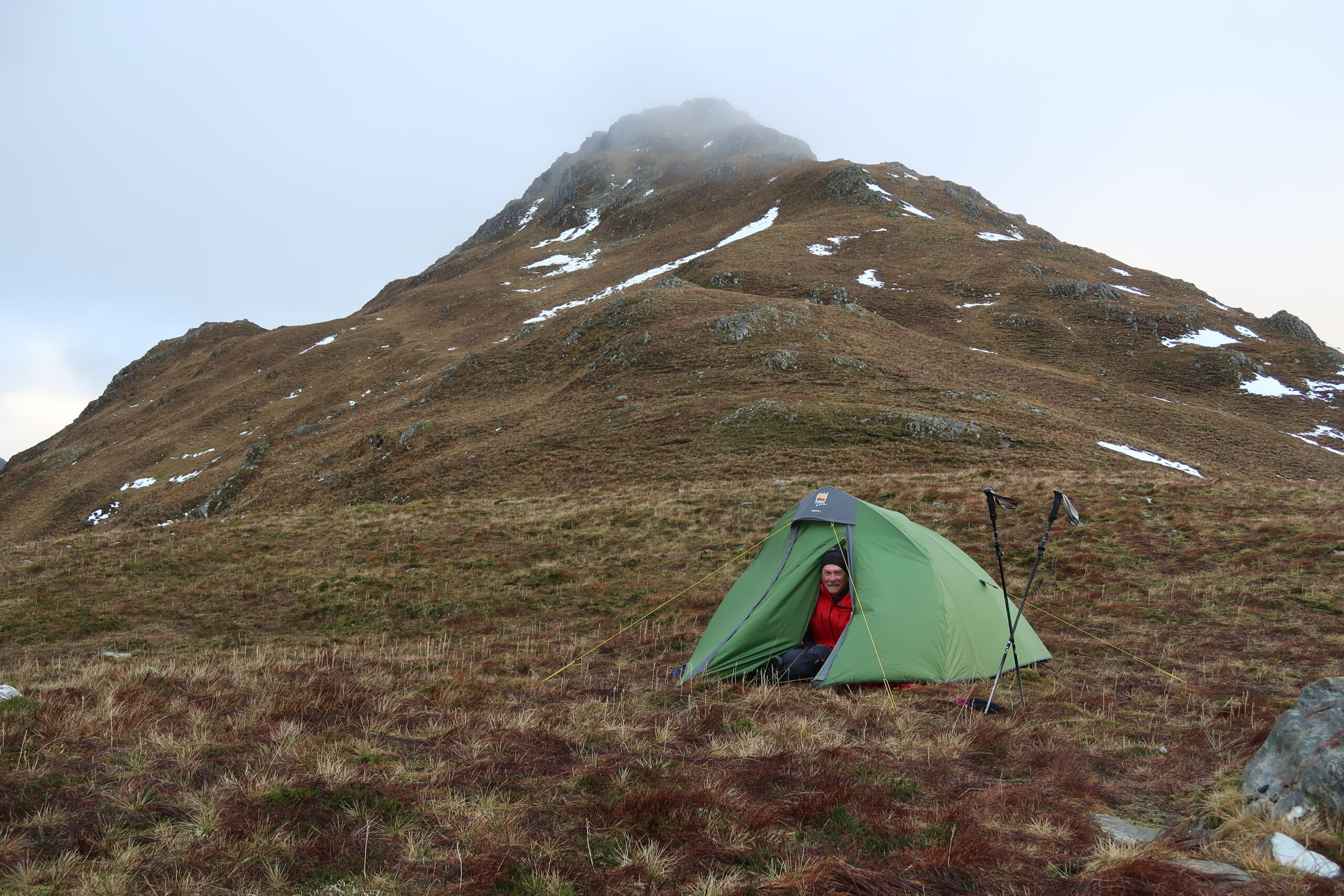 Person in a red jacket and beanie sits at the doorway of a green tent pitched on windswept brown grass with trekking poles beside it, set against a steep mist-capped mountain slope with patches of snow.