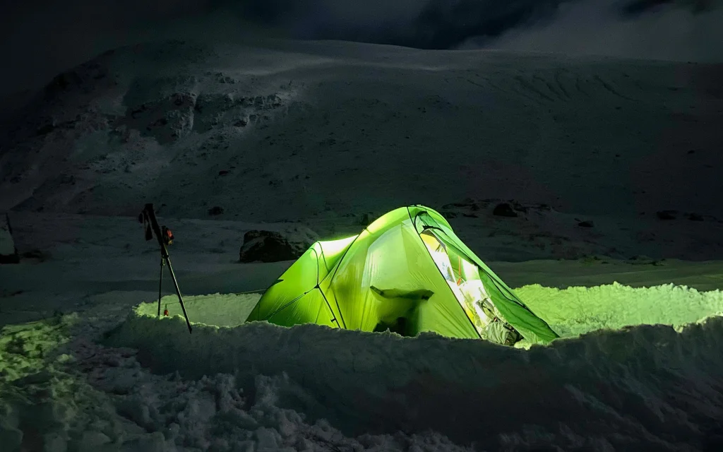 ls_night_snow_outdoorintrigue-2022 A glowing green tent illuminated from within sits in a dug-out snow trench on a dark, snow-covered mountain slope at night, with a set of trekking poles or a tripod standing nearby.