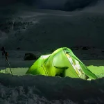 A glowing green tent illuminated from within sits in a dug-out snow trench on a dark, snow-covered mountain slope at night, with a set of trekking poles or a tripod standing nearby.