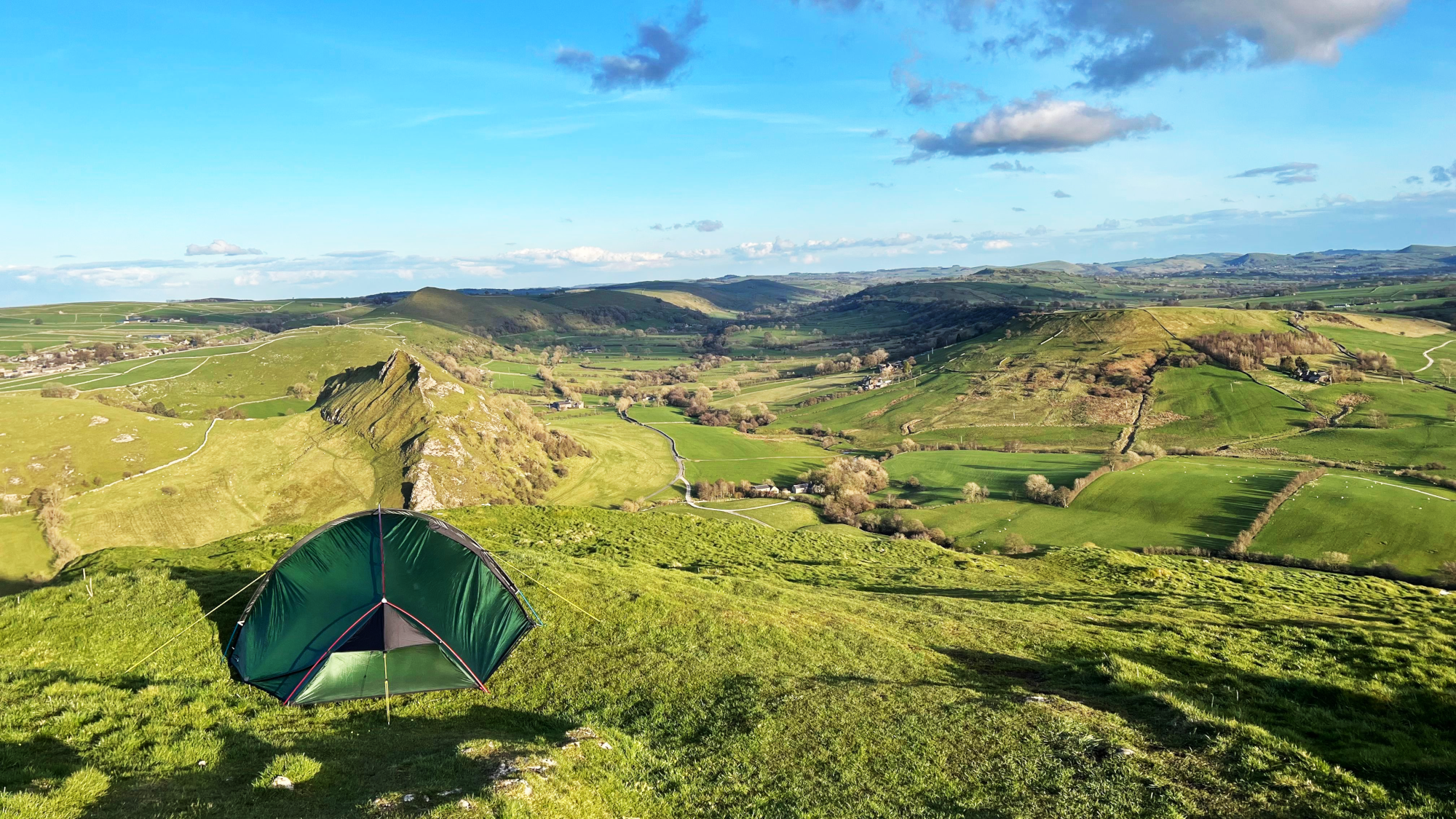 Small green tent pitched on a grassy hilltop in the foreground, overlooking a sweeping patchwork of rolling green fields, limestone ridges and scattered farmhouses under a bright blue sky with a few clouds.