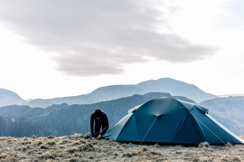 Person in dark clothing bending to secure a blue-green tent on a grassy mountain ridge with layered hills in the background under a pale, overcast sky