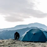 Person in dark clothing bending to secure a blue-green tent on a grassy mountain ridge with layered hills in the background under a pale, overcast sky