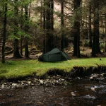 Green camping tent pitched on a grassy riverbank beside a rocky stream, set against a backdrop of tall, dense conifer trees.