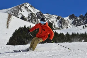 A skier in an orange jacket, tan trousers, black helmet and reflective goggles carves through snow, sending up a spray while holding poles against a backdrop of snow‑covered mountains and a clear blue sky.