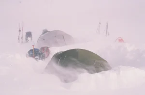 Small dome tents and a larger tent partly buried in deep snow at a wind-blown alpine campsite, with skis, sledges and backpacks standing nearby in near-whiteout conditions