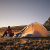 ls_solar_ultra_2_mountian_2023 Camper in a red jacket sits on a grassy alpine plateau at sunset, unpacking gear from a foil container beside a pitched orange tent with rocky mountains in the background.