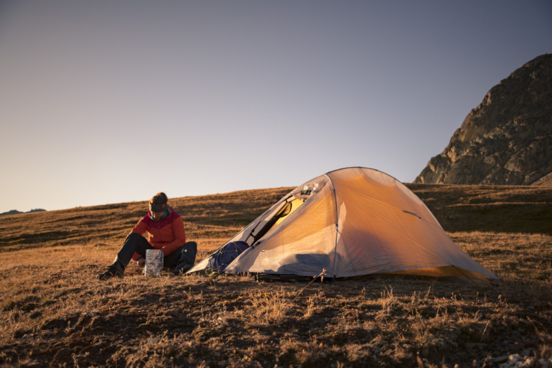 ls_solar_ultra_2_mountian_2023 Camper in a red jacket sits on a grassy alpine plateau at sunset, unpacking gear from a foil container beside a pitched orange tent with rocky mountains in the background.