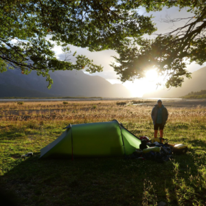 A green one‑person tent pitched under leafy tree branches on sunlit grass beside a wide river plain, a person standing near piled camping gear as the sun rises behind distant mountains.