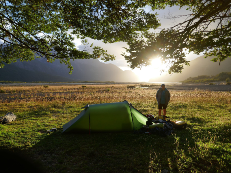 ls_starlite_2_2019sa45-1-scaled A green one‑person tent pitched under leafy tree branches on sunlit grass beside a wide river plain, a person standing near piled camping gear as the sun rises behind distant mountains.