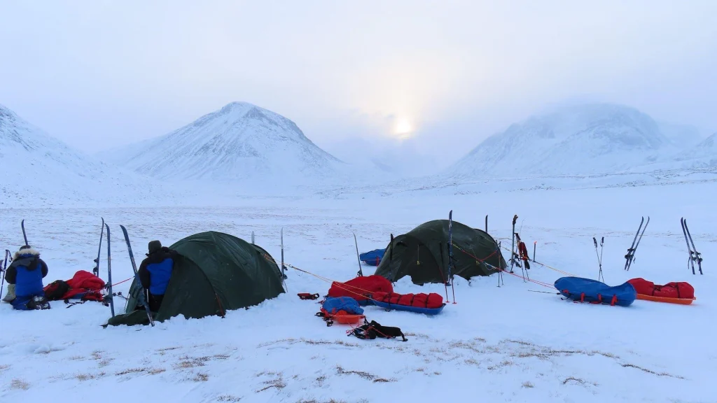 Two green expedition tents pitched on a snowy plain in a mountain valley at dawn, surrounded by skis, sleds and red and blue gear with two bundled figures working beside the tents and pale sun breaking through clouds above snow‑covered peaks.
