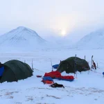 Two green expedition tents pitched on a snowy plain in a mountain valley at dawn, surrounded by skis, sleds and red and blue gear with two bundled figures working beside the tents and pale sun breaking through clouds above snow‑covered peaks.