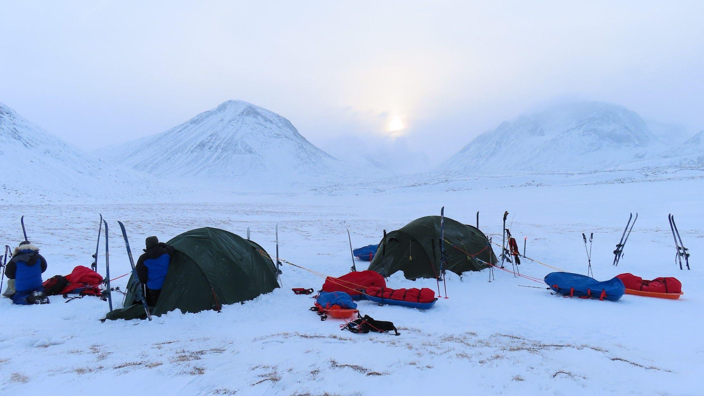 Two green expedition tents pitched on a snowy plain in a mountain valley at dawn, surrounded by skis, sleds and red and blue gear with two bundled figures working beside the tents and pale sun breaking through clouds above snow‑covered peaks.
