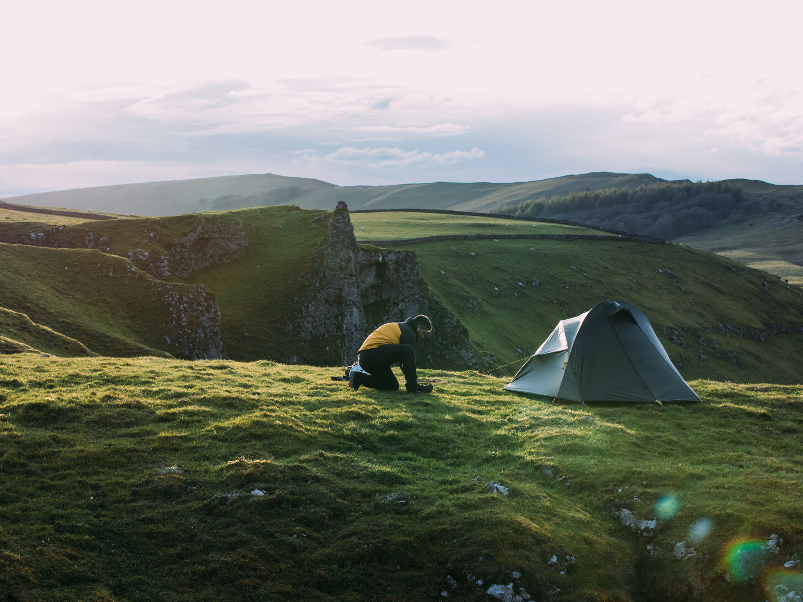 A camper kneels in a yellow jacket beside a small grey tent on a grassy cliff-top, overlooking rolling green hills under a pale sky.