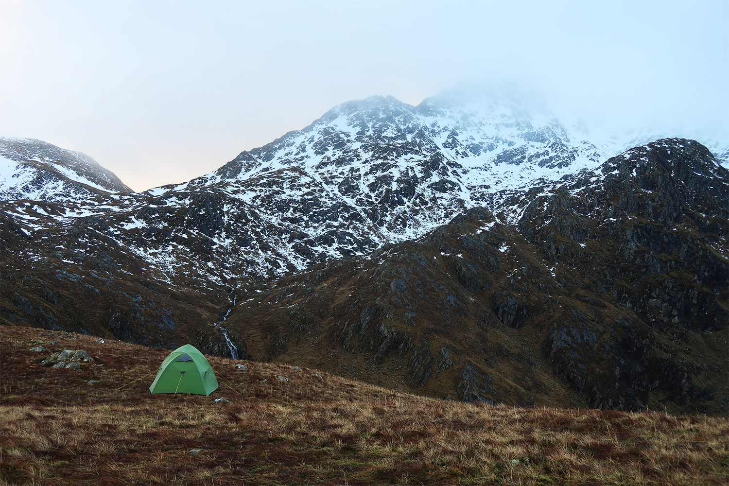 A solitary green tent on a brown grassy slope in the foreground with steep, snow-speckled rocky mountains and mist-shrouded peaks rising behind and a narrow stream running through the valley.