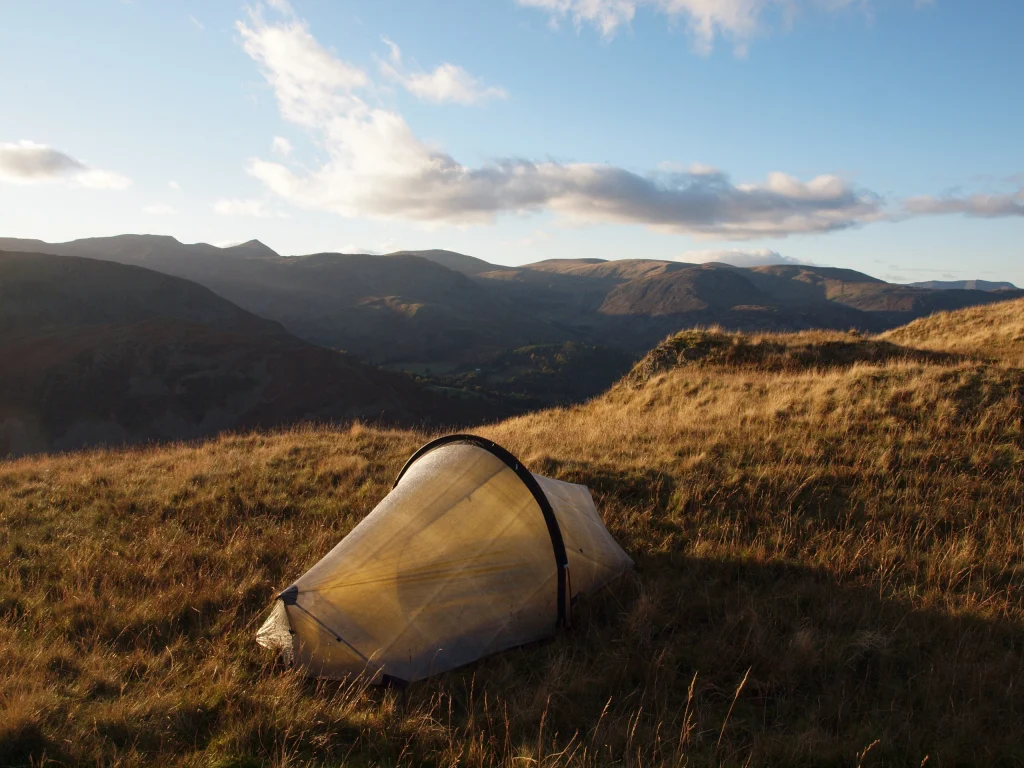A small translucent backpacking tent pitched on a sunlit grassy hillside, overlooking rolling mountains under a blue sky with scattered clouds.