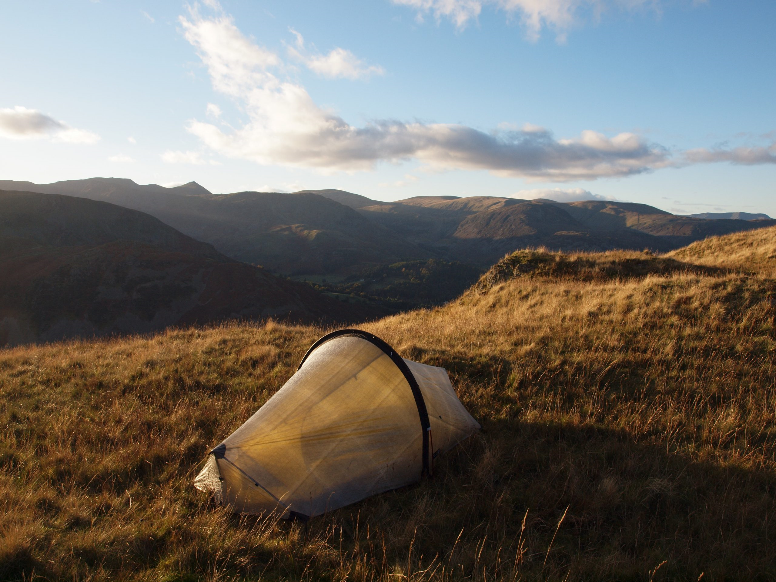 A small translucent backpacking tent pitched on a sunlit grassy hillside, overlooking rolling mountains under a blue sky with scattered clouds.