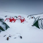 Several red and green dome tents, mostly covered in fresh snow and anchored with guy lines, sit in a snowy mountain campsite under a grey sky, one tent doorway showing a person in an orange hood.