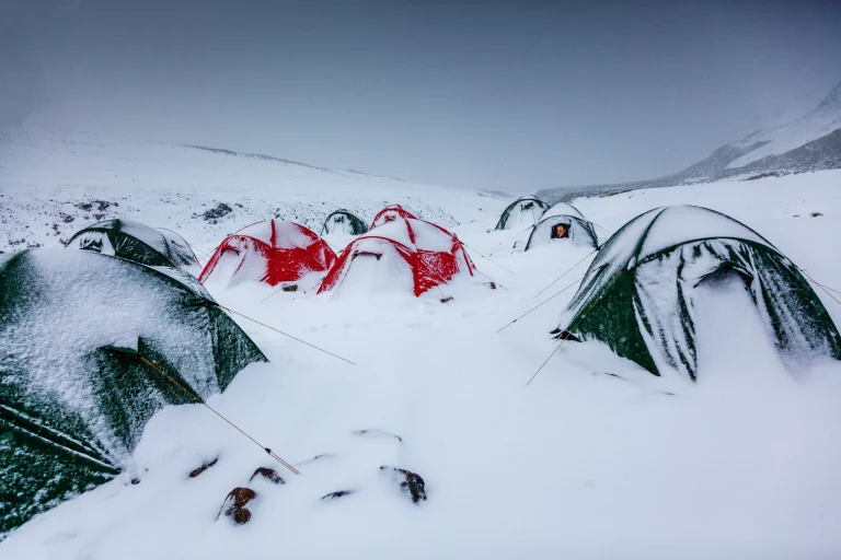 Several red and green dome tents, mostly covered in fresh snow and anchored with guy lines, sit in a snowy mountain campsite under a grey sky, one tent doorway showing a person in an orange hood.