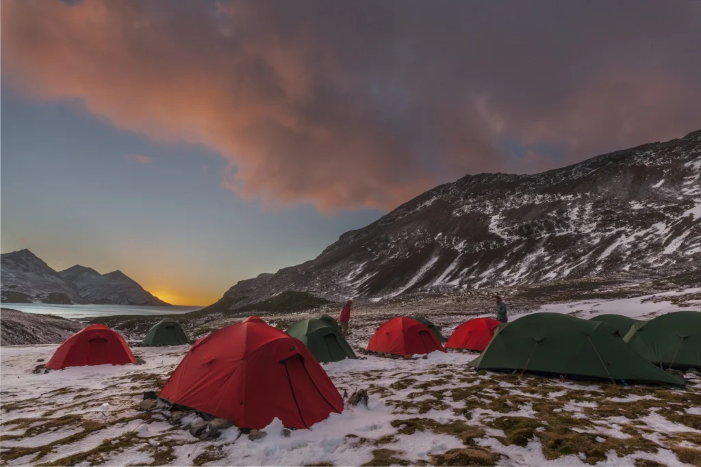 Red and green tents dot a snowy mountain campsite on a grass-and-snow plateau at sunset, beneath dramatic pink clouds with steep, snow-dusted ridges to the right and a sunlit fjord between distant peaks to the left.