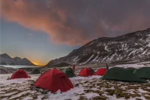 Two green dome tents pitched on a windswept grassy plateau beside a steep layered canyon, a camper sitting on the ground among gear with rolling hills and a bright blue sky in the background.