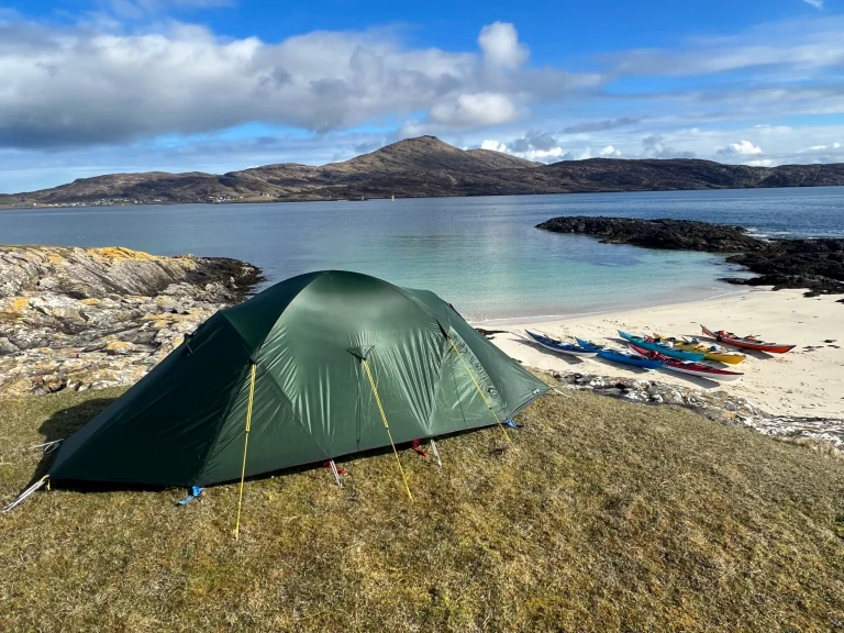 Dome-style green tent pitched on a grassy headland above a white sandy beach lined with colourful kayaks, clear turquoise water, rocky outcrops and rolling hills under a blue sky with scattered clouds