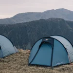 Three pale-blue dome tents staked on a windswept grassy ridge beneath steep, rocky mountains under a pale overcast sky.