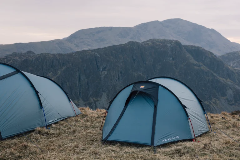 Three pale-blue dome tents staked on a windswept grassy ridge beneath steep, rocky mountains under a pale overcast sky.
