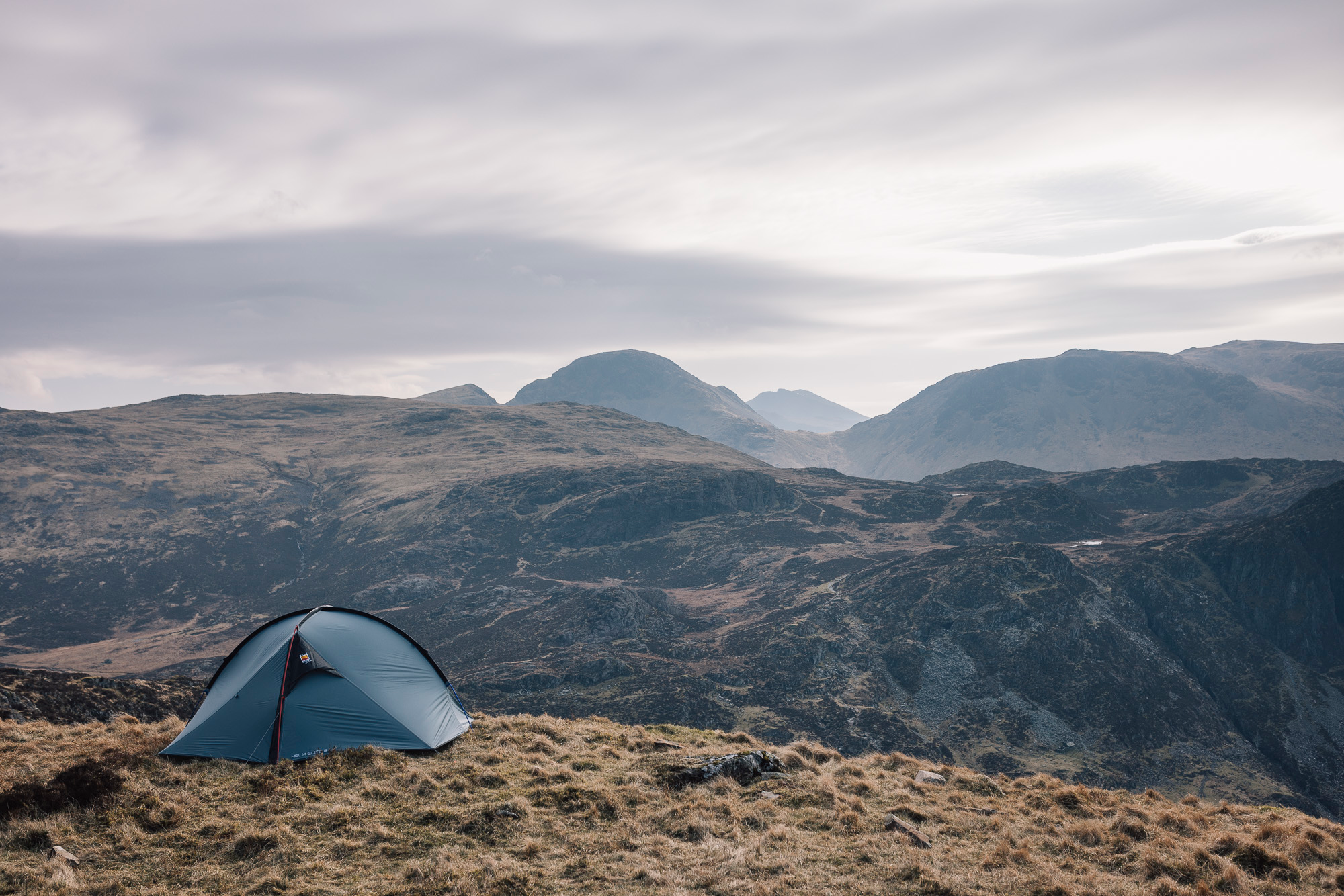 A small blue two-person tent pitched on windswept grassy moorland overlooking layered rugged mountains under a pale, cloudy sky.