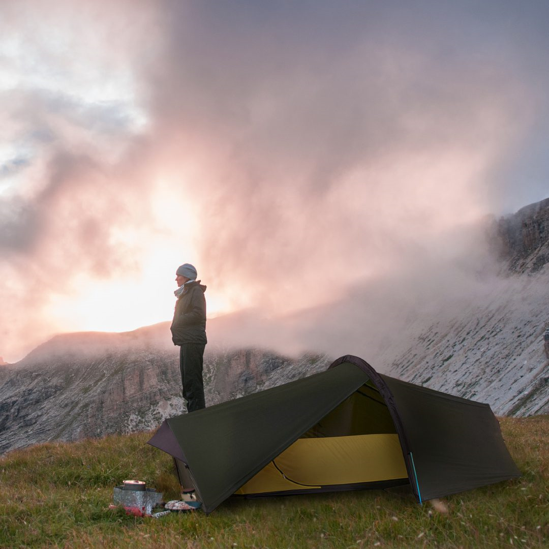 A camper wearing a beanie and jacket stands beside a low-profile green and yellow tent on a grassy mountain ridge as pink dawn clouds glow above rocky cliffs, with a small cooking pot on the ground nearby.