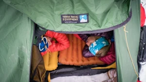 ls_tn_logo_2024 Top-down view into a green Terra Nova tent with its logo patch visible, showing two people in winter jackets lying on orange and yellow sleeping mats—one wearing a blue patterned headband—with snow visible outside.