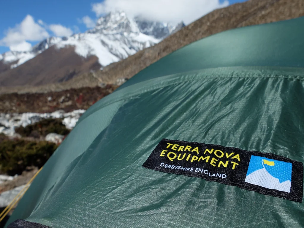 ls_tn_logo_close_up_mountian_2021 Green tent flysheet with a prominent 'Terra Nova Equipment, Derbyshire England' stitched label in the foreground and snow‑capped mountains and rocky alpine terrain blurred in the background.