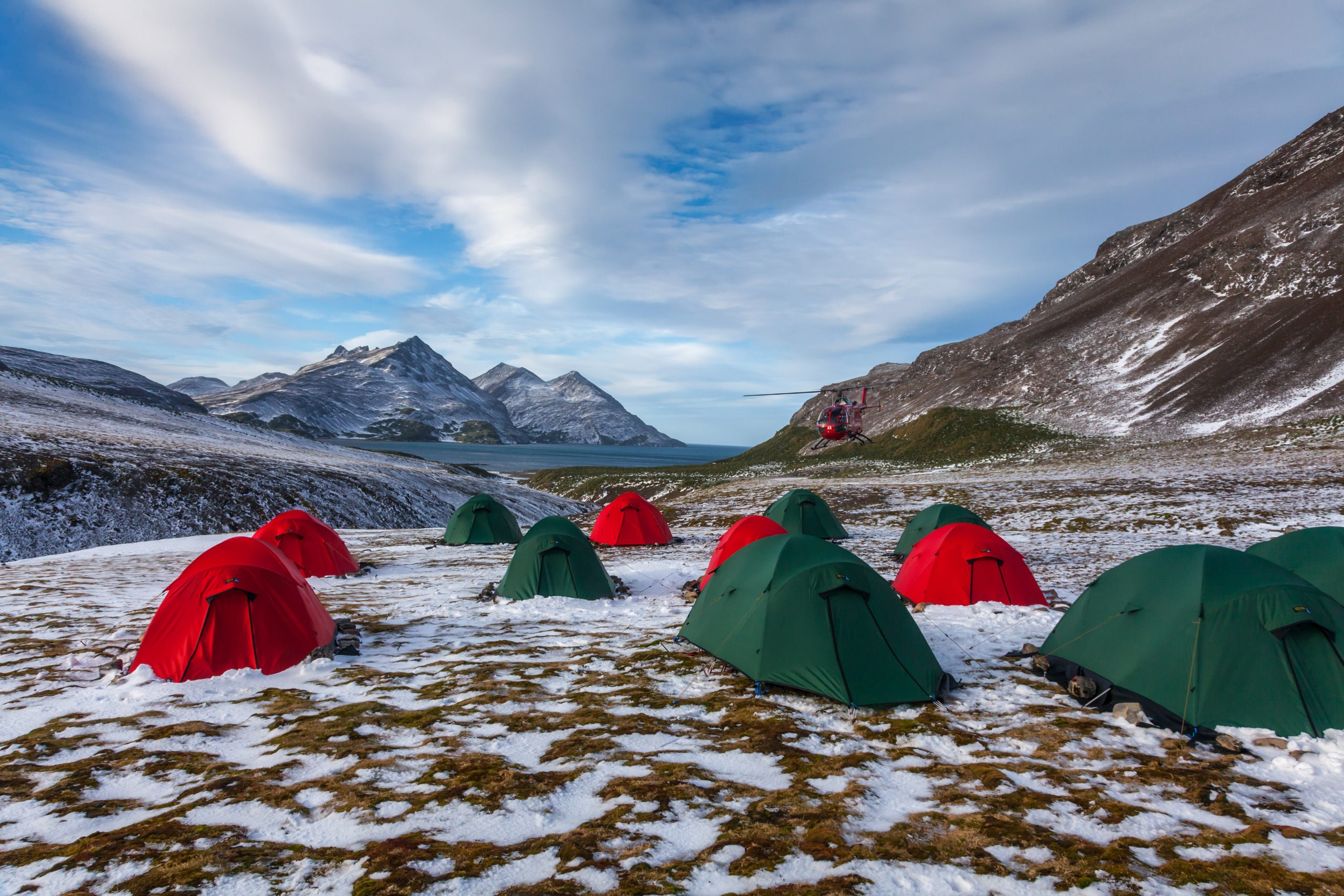 Cluster of red and green dome tents pitched on patchy snow-covered tundra with rugged, snow-dusted mountains and a distant fjord under a cloudy sky, a red helicopter hovering near a hillside.