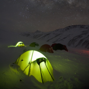 Two green dome tents pitched on a windswept grassy plateau beside a steep layered canyon, a camper sitting on the ground among gear with rolling hills and a bright blue sky in the background.