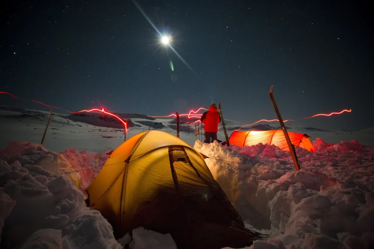 ls_tn_night_snow_quasar-tent Night-time snowy mountain campsite under a bright moon and starry sky with a glowing yellow tent in the foreground, an illuminated orange tent and a person in a red jacket standing by upright skis amid snow trenches, while red headlamp light trails streak across the scene.