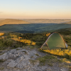 ls_tn_peaks_Southern Cross_2021 Green one-person tent pitched on a rocky, grassy hilltop at golden hour, overlooking rolling valleys, distant hills and a sunlit reservoir.