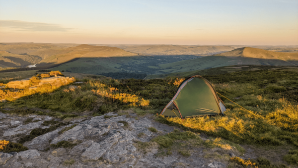 ls_tn_peaks_Southern Cross_2021 Green one-person tent pitched on a rocky, grassy hilltop at golden hour, overlooking rolling valleys, distant hills and a sunlit reservoir.