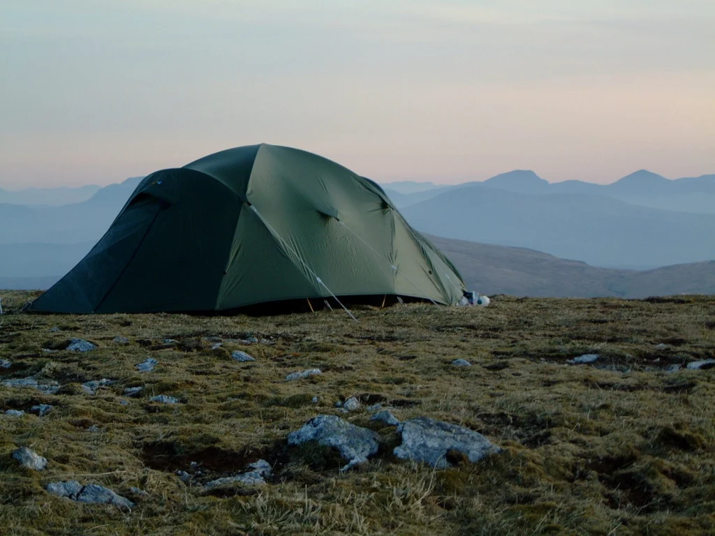 ls_tn_quasar_2023 A green dome-style camping tent staked on a rocky, grassy upland at dusk with pale sky and layered blue mountain ridges in the distance.