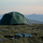 A green dome-style camping tent staked on a rocky, grassy upland at dusk with pale sky and layered blue mountain ridges in the distance.