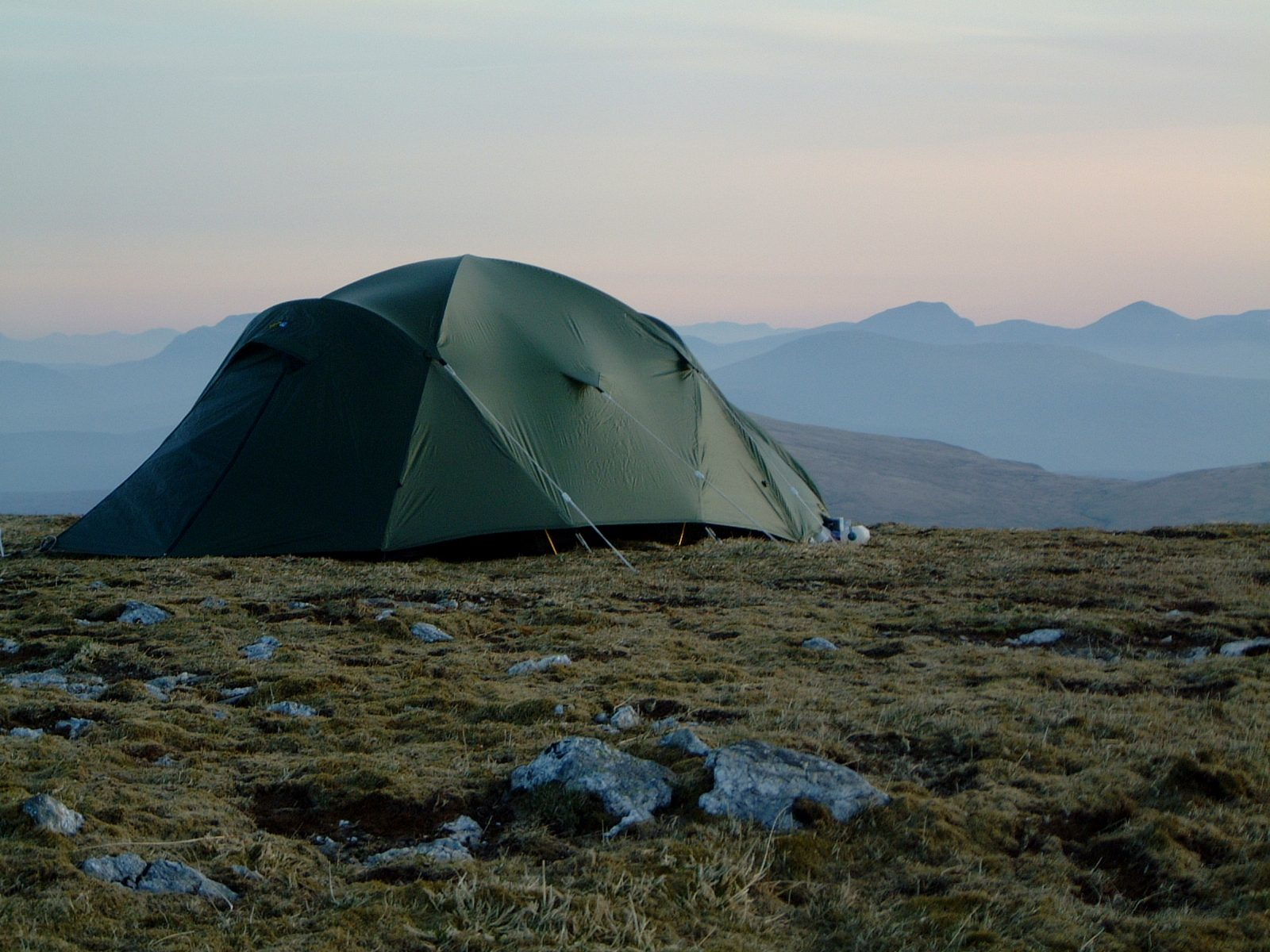 A green dome-style camping tent staked on a rocky, grassy upland at dusk with pale sky and layered blue mountain ridges in the distance.