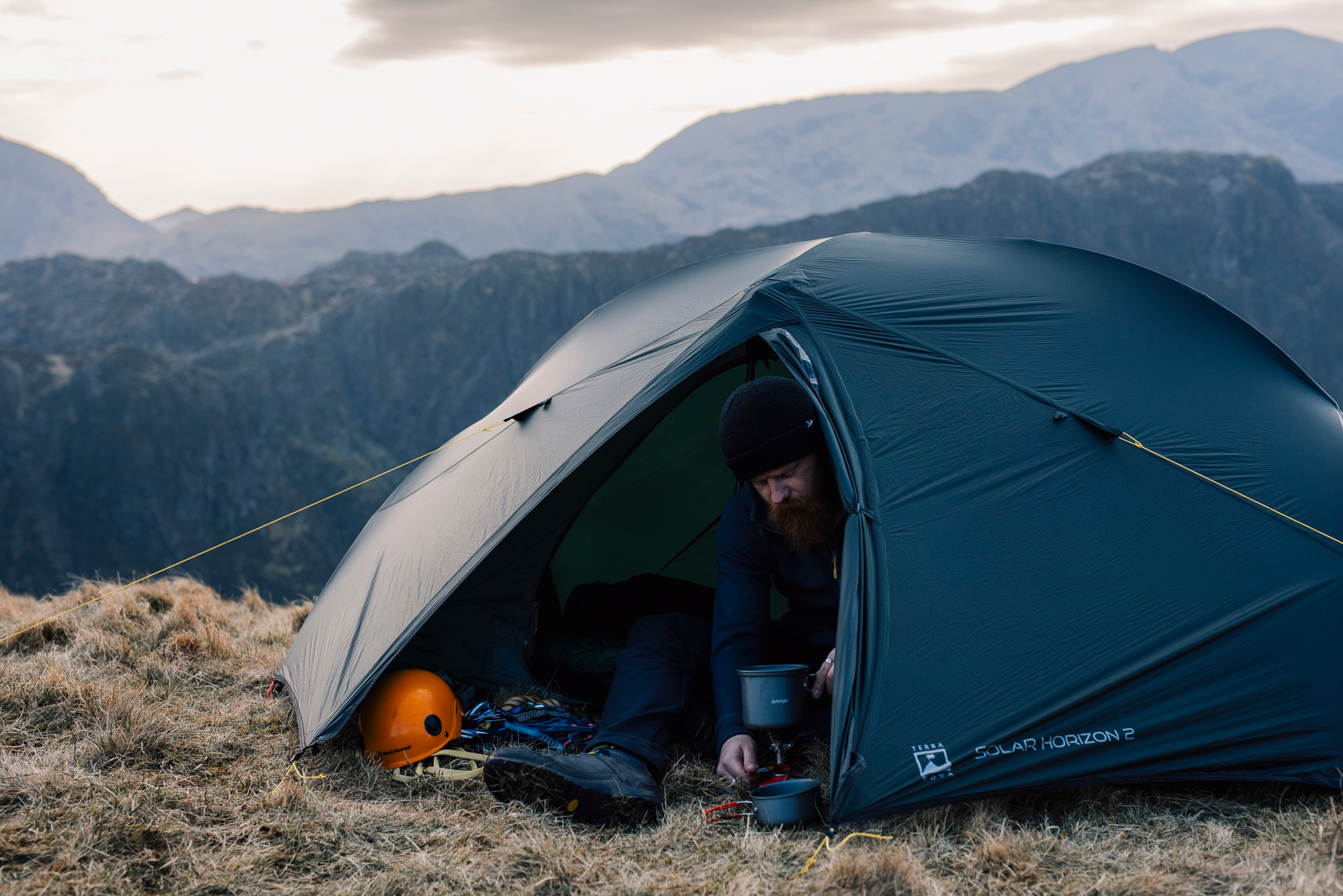 A green dome-style camping tent staked on a rocky, grassy upland at dusk with pale sky and layered blue mountain ridges in the distance.