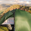 ls_tn_southern_cross__2021 Green backpacking tent pitched on a grassy mountain ridge with red poles and a steaming pot visible inside, overlooking layered rocky peaks under a partly cloudy sky.