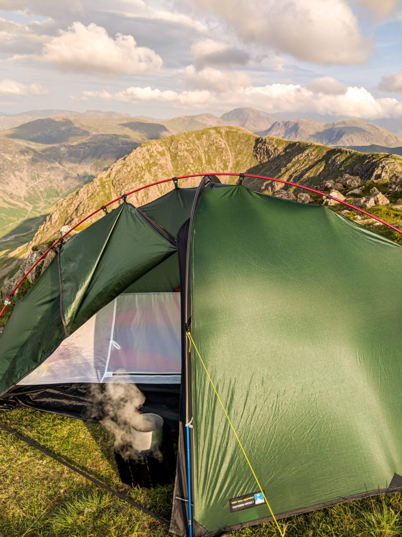 ls_tn_southern_cross__2021 Green backpacking tent pitched on a grassy mountain ridge with red poles and a steaming pot visible inside, overlooking layered rocky peaks under a partly cloudy sky.