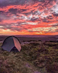 A small dome tent pitched on grassy moorland beneath a dramatic pink and orange sunset sky with layered clouds stretching over distant rolling hills.