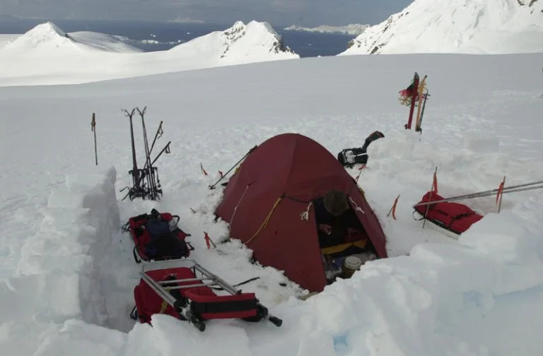 Red dome tent pitched in a dug-out snow shelter surrounded by sleds, skis, poles and gear on a vast snowy mountain plateau with distant snow-covered peaks.