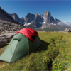 ls_tn_voyager-Harsharn-Gill_2021 Green backpacking tent with a red sleeping bag draped over it, pitched on a grassy alpine meadow dotted with yellow wildflowers and rocky outcrops, with steep jagged snow-patched mountains and a clear blue sky in the background.