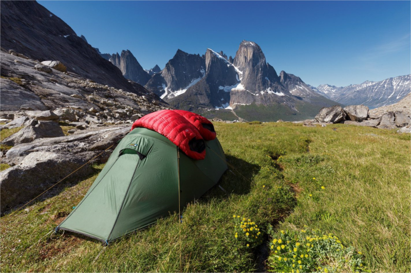 Green backpacking tent with a red sleeping bag draped over it, pitched on a grassy alpine meadow dotted with yellow wildflowers and rocky outcrops, with steep jagged snow-patched mountains and a clear blue sky in the background.