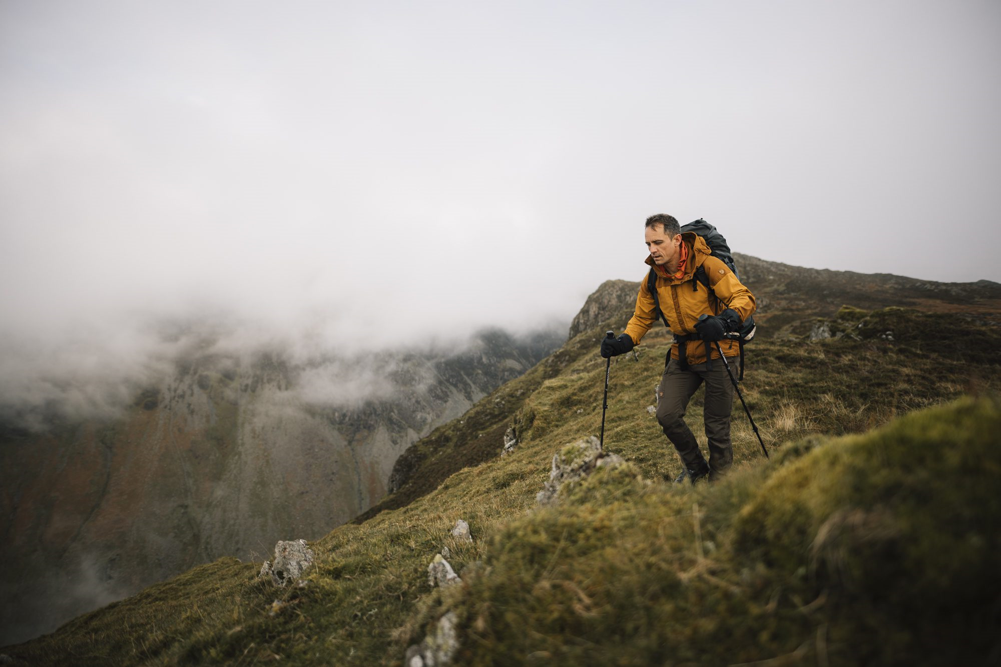 A hiker wearing a yellow jacket and backpack uses trekking poles while ascending a grassy, fog‑shrouded mountain ridge.
