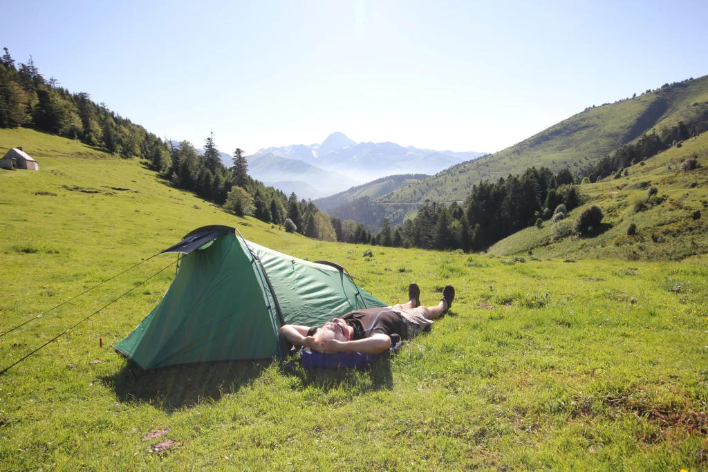 Camper lying on their back with hands behind their head beside a small green tent on a sunlit grassy mountain meadow, with tree-covered hills and distant snow-capped peaks under a clear sky.